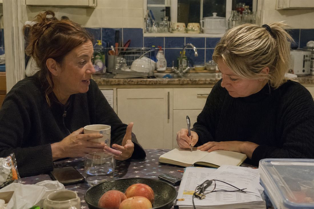 Two women at a kitchen table, one talking while the other takes notes
