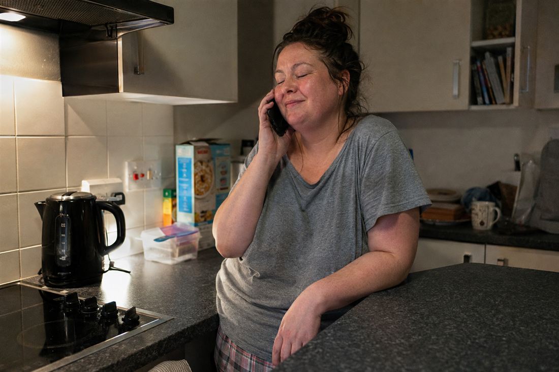 A woman leaning on her kitchen counter, eyes closed with relief on a phone call